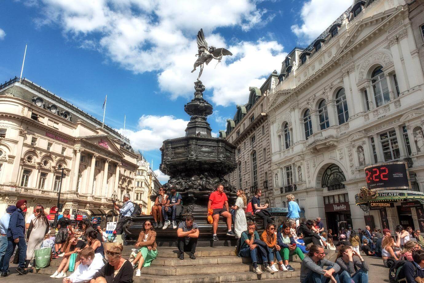 Tourists sitting on the steps of the Shaftesbury Memorial Fountain and statue of Greek god Anteros, Piccadilly Circus, London’s West End.